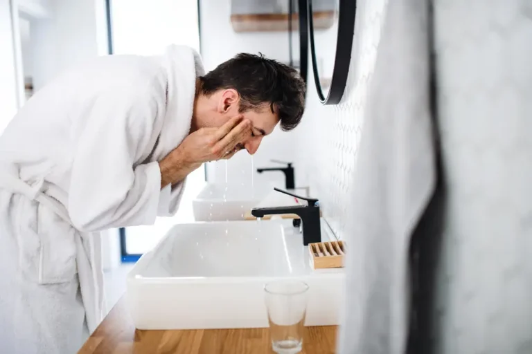 A man in a white bathrobe washing his face at a modern bathroom sink, following an acne treatment skincare routine