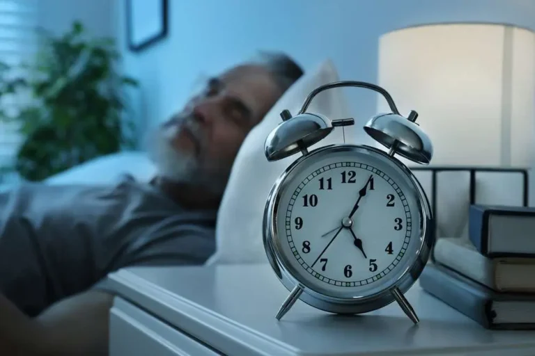 A close-up of a silver alarm clock on a nightstand, showing 1:55 AM, with an elderly man sleeping in the background, representing the importance of Sleep and Fatigue Treatment for better rest